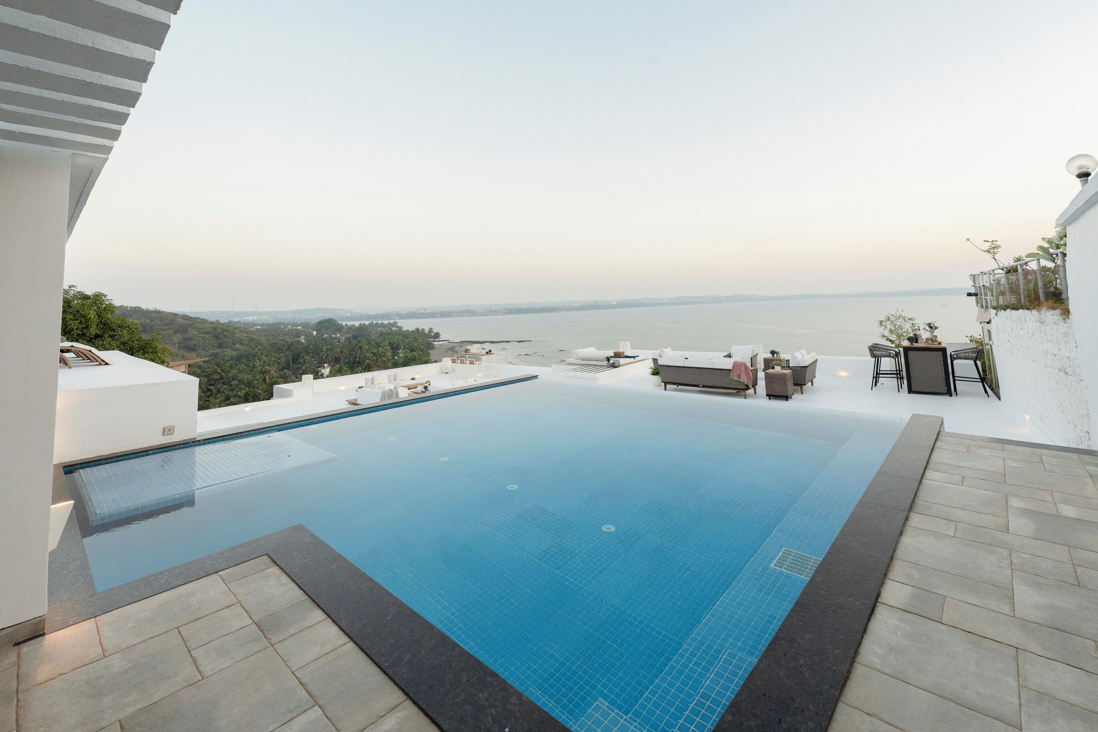 A swimming pool with a view of the sea, outdoor seating, and a white wall.
