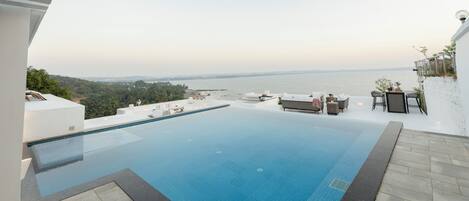 A swimming pool with a view of the sea, outdoor seating, and a white wall.