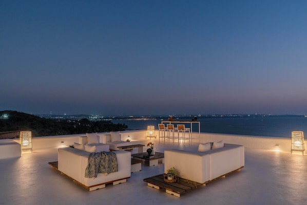 A rooftop terrace with white sofas, a wooden table, and a bar area.