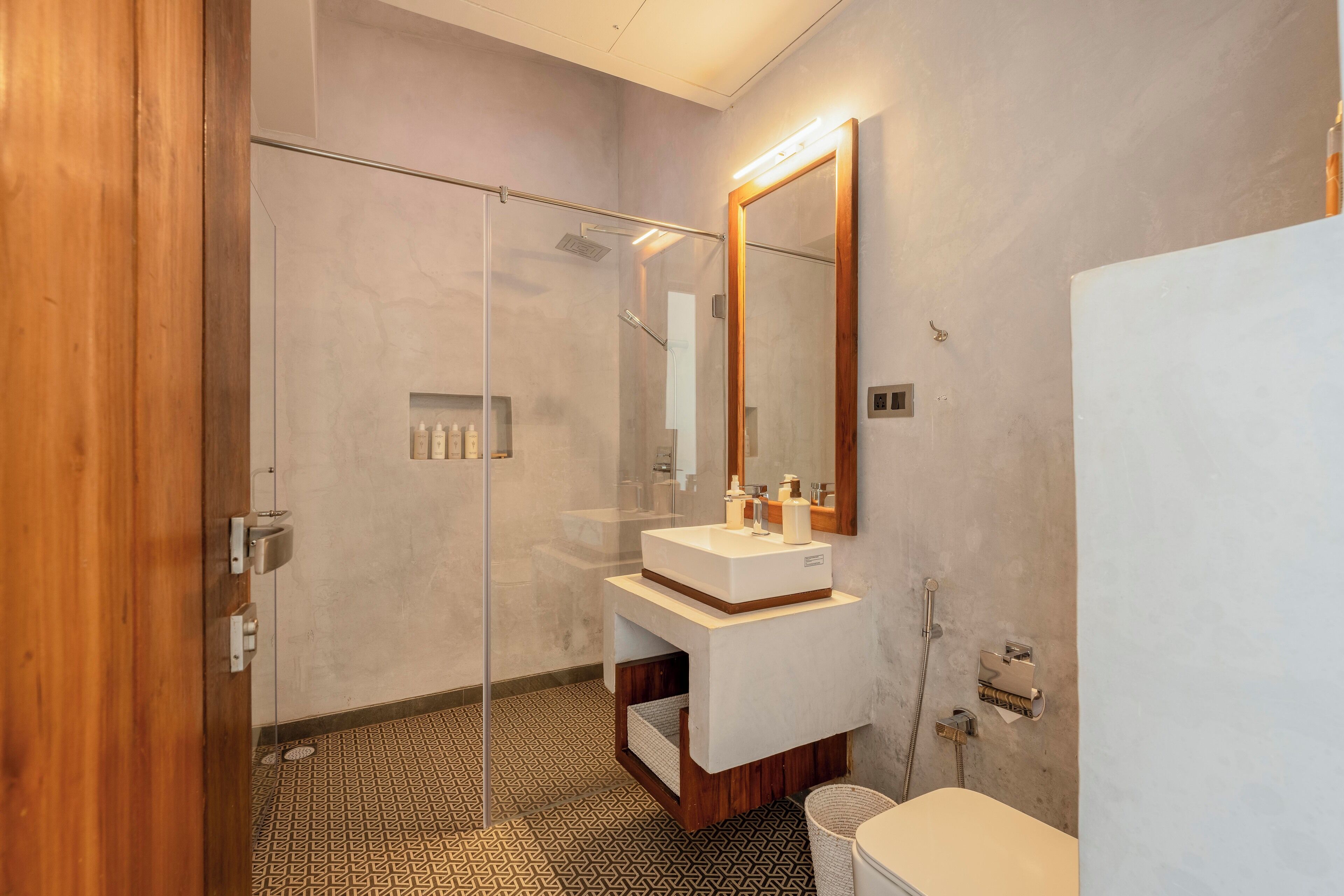 A modern bathroom with a glass shower enclosure, a wooden door, a white sink with a mirror, and patterned floor tiles.