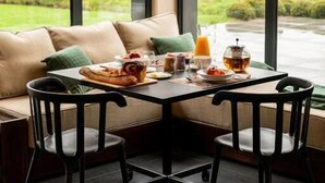 Un coin repas avec une table mise pour le petit-déjeuner, un canapé et de grandes fenêtres donnant sur un paysage verdoyant.