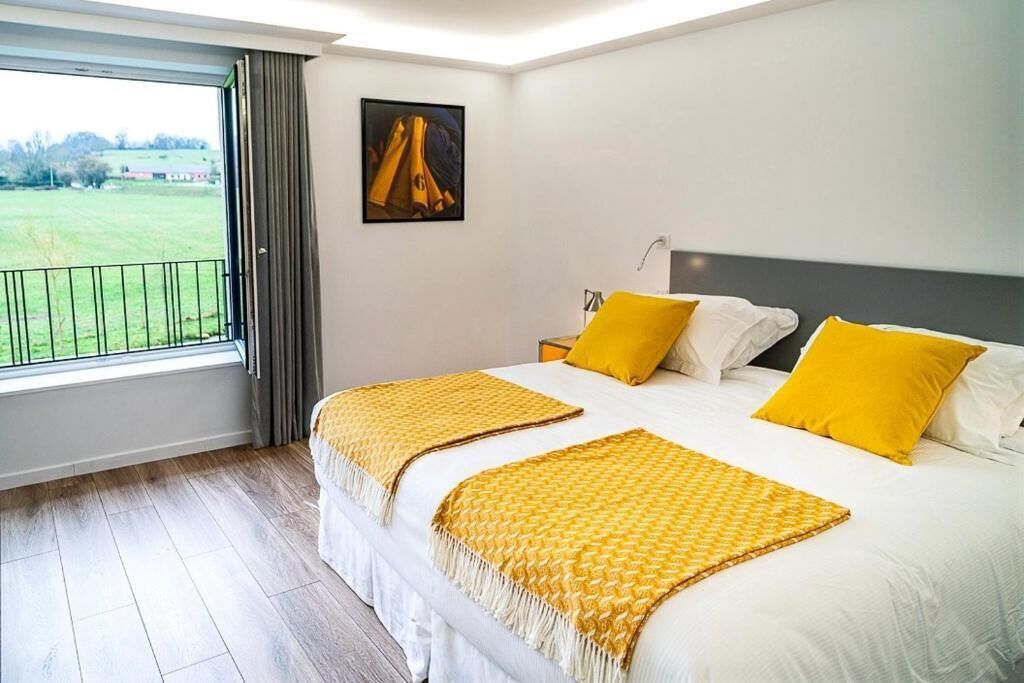 Hotelkamer met een groot bed, twee gele kussens, een witte spreideken, een geel plaid, een schilderij aan de muur en uitzicht op een groen veld door een raam.