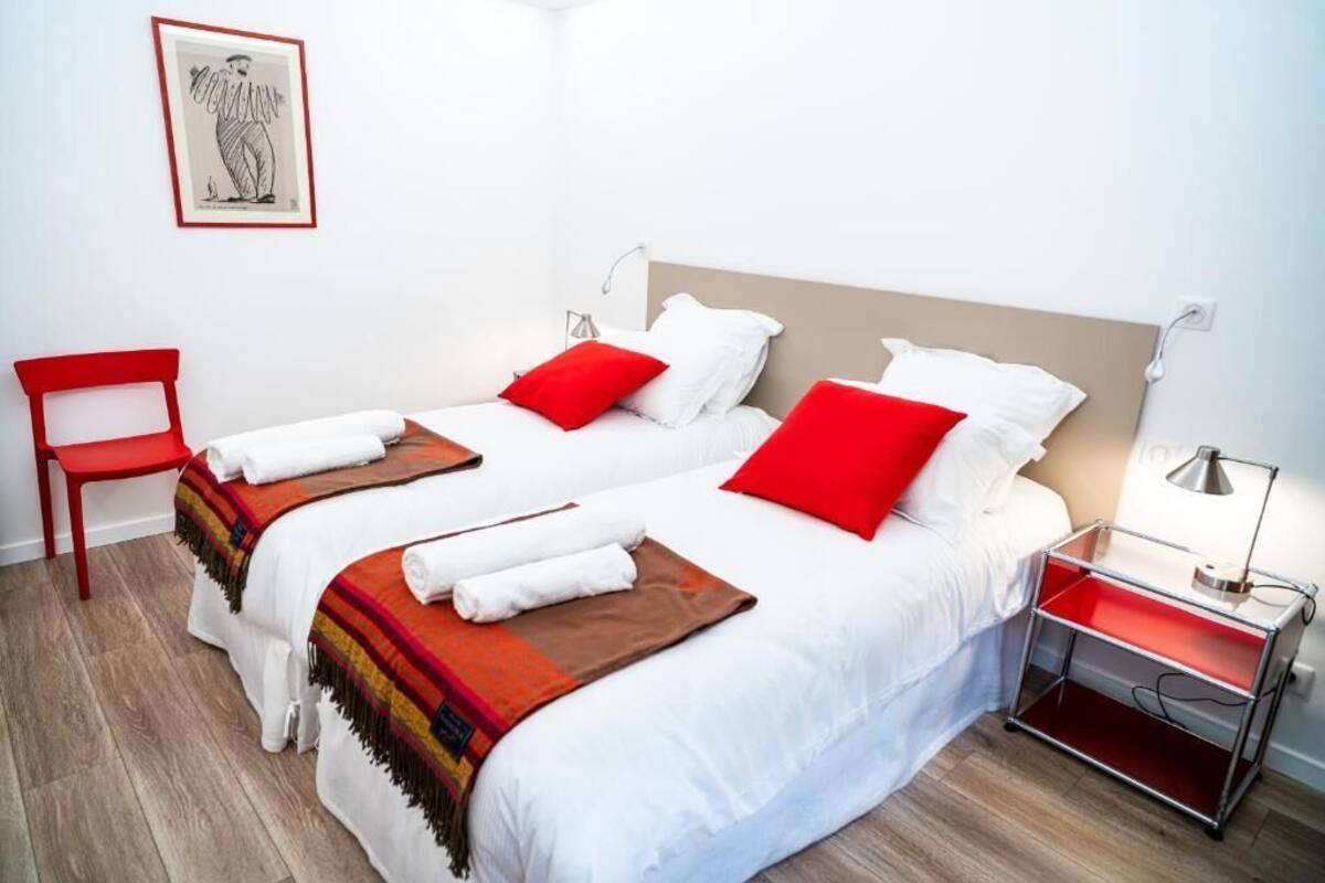A hotel room with two beds, red pillows, a red chair, a nightstand with a lamp, and a framed picture on the wall.