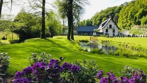 Un bâtiment blanc avec un toit foncé, entouré d’une pelouse bien entretenue et d’un petit étang.
