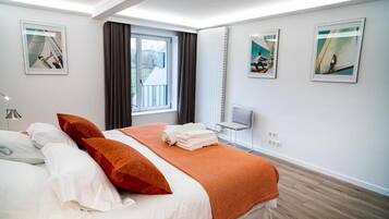 Ett modernt hotellrum med en stor säng, ett fönster med gardiner, en stol och konstverk på väggarna.