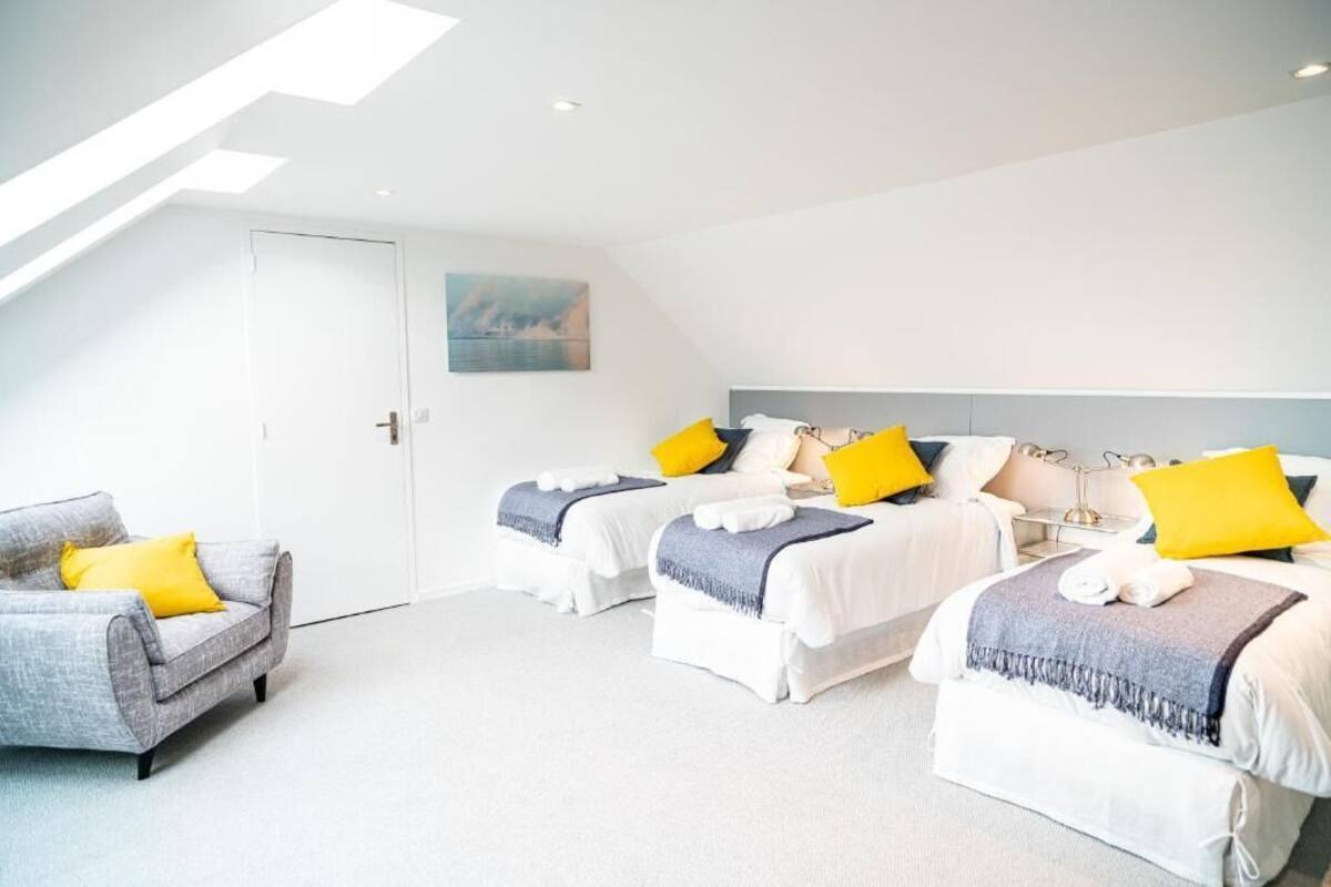A modern hotel room with a loft-style ceiling, two beds with matching headboards, a gray sofa with yellow pillows, and a painting on the wall.