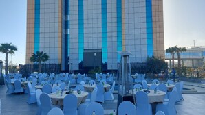 An outdoor dining area with round tables and white chairs, set against a modern building with blue and beige accents. - Taj Al- Wajh hotel   (Al Wajh)