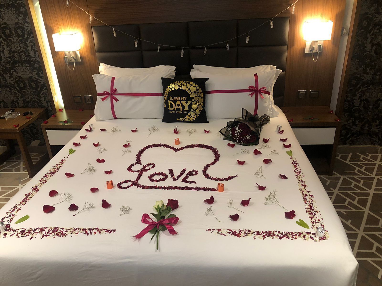Cama bem arrumada com decoração em formato de coração com a palavra 'LOVE', pétalas de rosa e um buquê de flores.