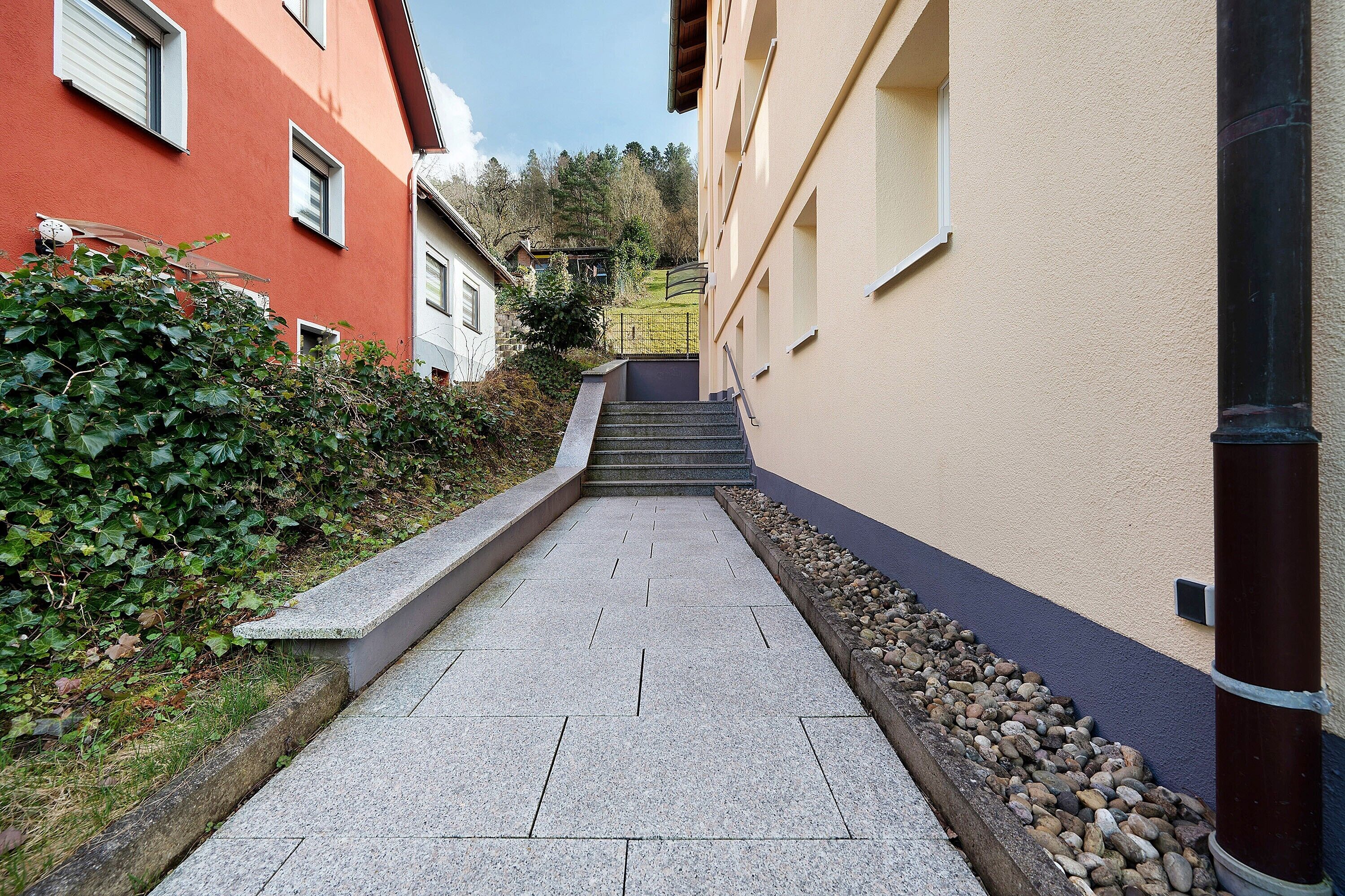Ein schmaler, gepflasterter Weg, der von Gebäuden gesäumt wird, mit einer Steinsitzbank und einem Metallgeländer.