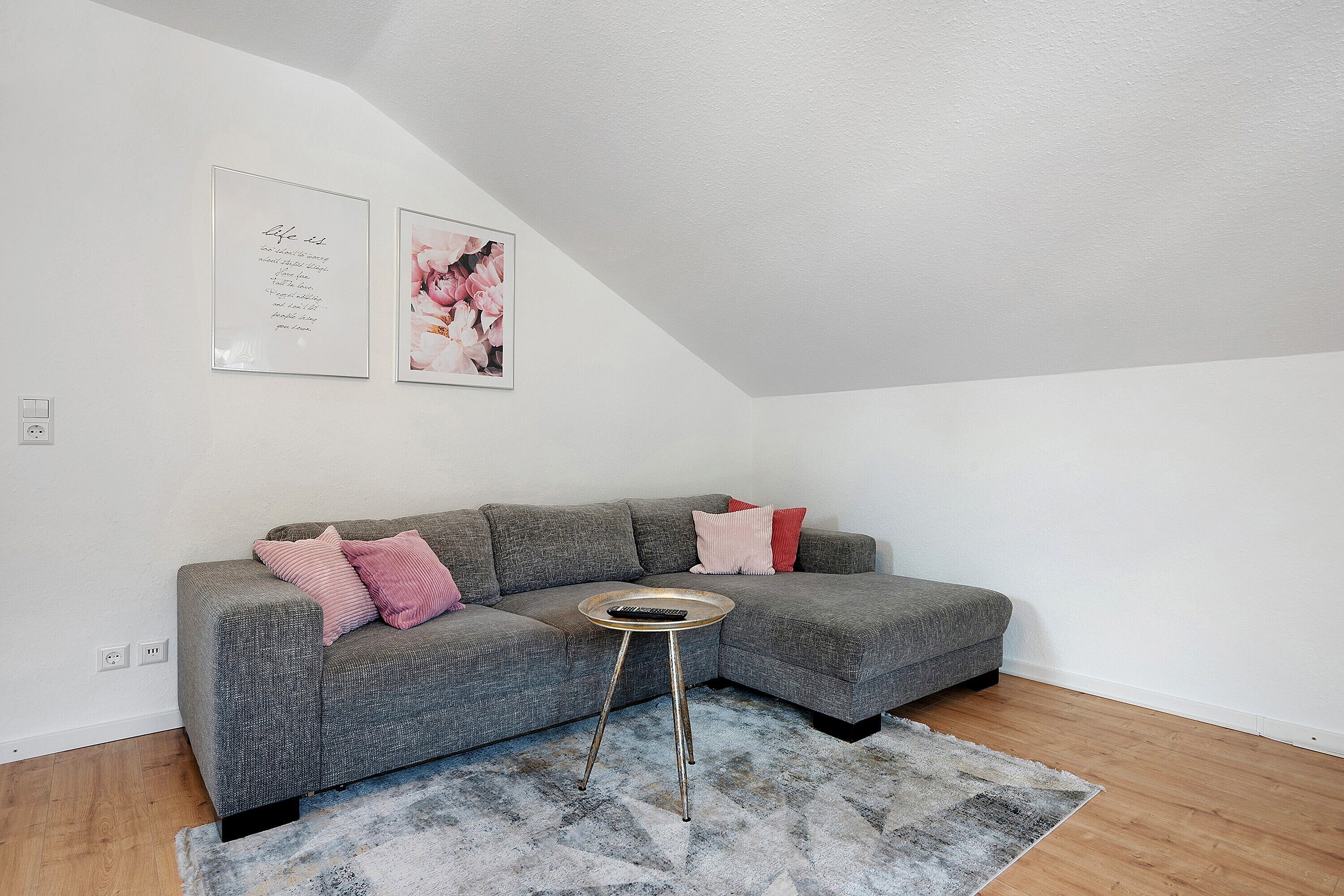 A gray sectional sofa with pink and red pillows, a small round table with a remote control, and a gray rug on a wooden floor.