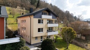A multi-story building with a balcony on the second floor, a visible number '100' on the facade, and a surrounding landscape with trees and a hill.