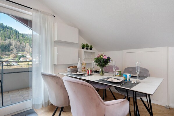 A dining area with a table set for two, a view of a balcony, and a kitchen area with cabinets and a countertop.