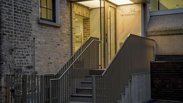Un escalier menant à l’entrée d’un immeuble, avec une plaque portant le numéro « 94 », un mur de briques et une rambarde en métal.