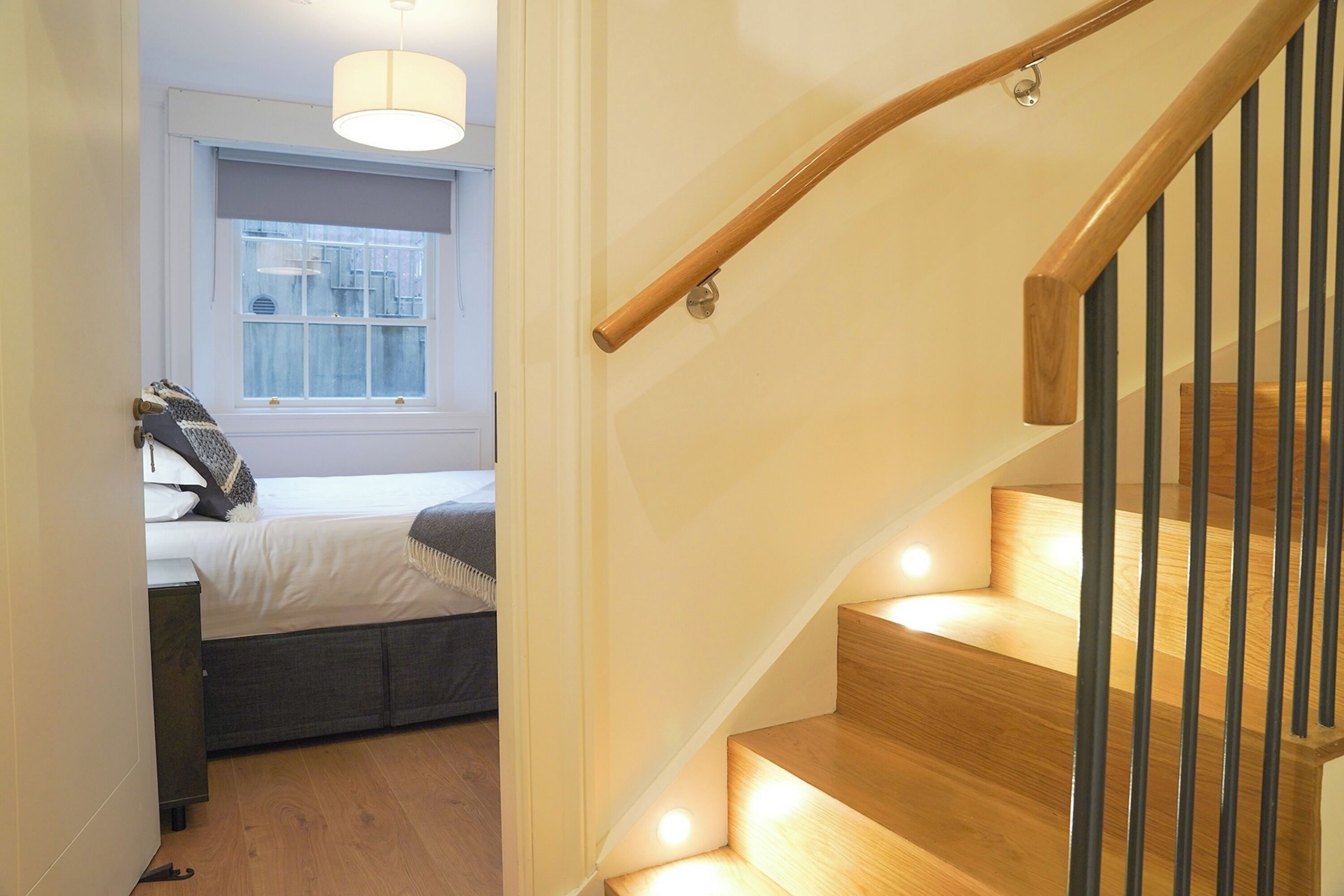 A staircase with wooden steps and black railings, leading to a bedroom with a bed and a window.