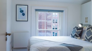Une chambre avec un grand lit, une fenêtre donnant sur des immeubles, un radiateur et un tableau encadré accroché au mur.