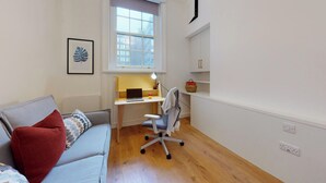 Un espace de vie compact comprenant un canapé, un bureau avec un ordinateur portable, une chaise et une fenêtre donnant sur des immeubles.