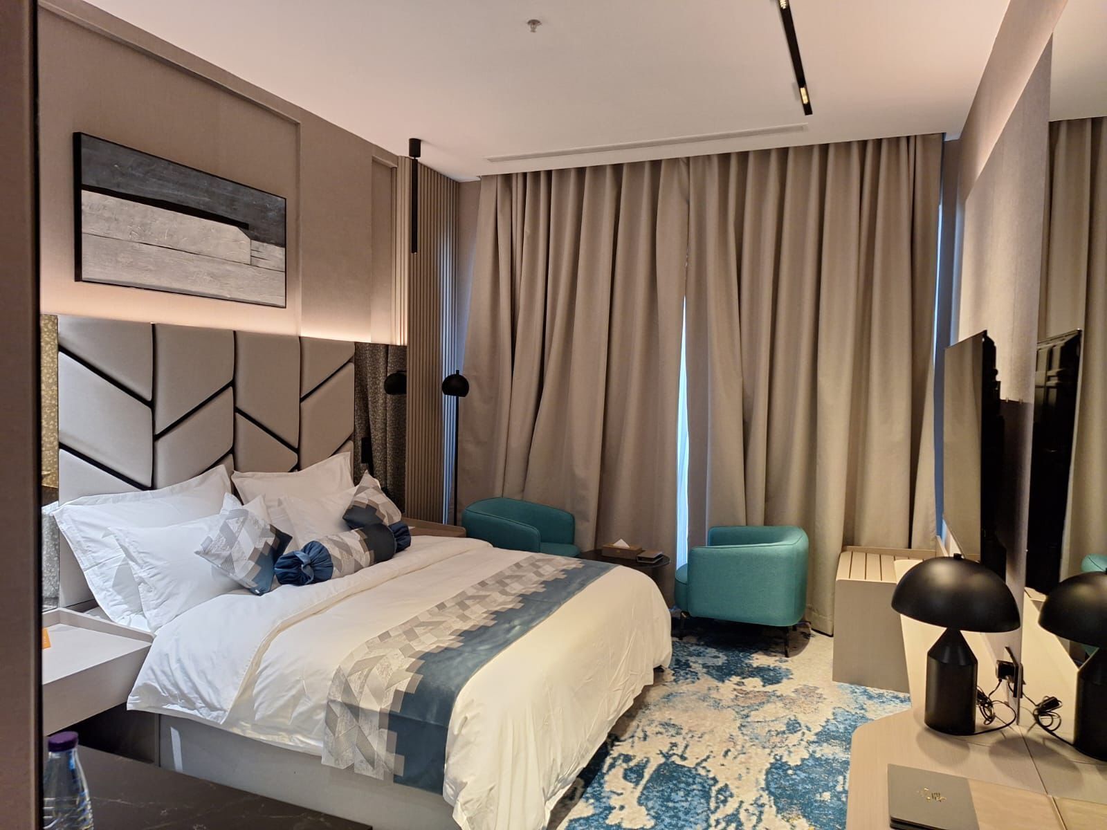 A modern hotel room with a large bed, a desk, a chair, a TV, and a large window with curtains.