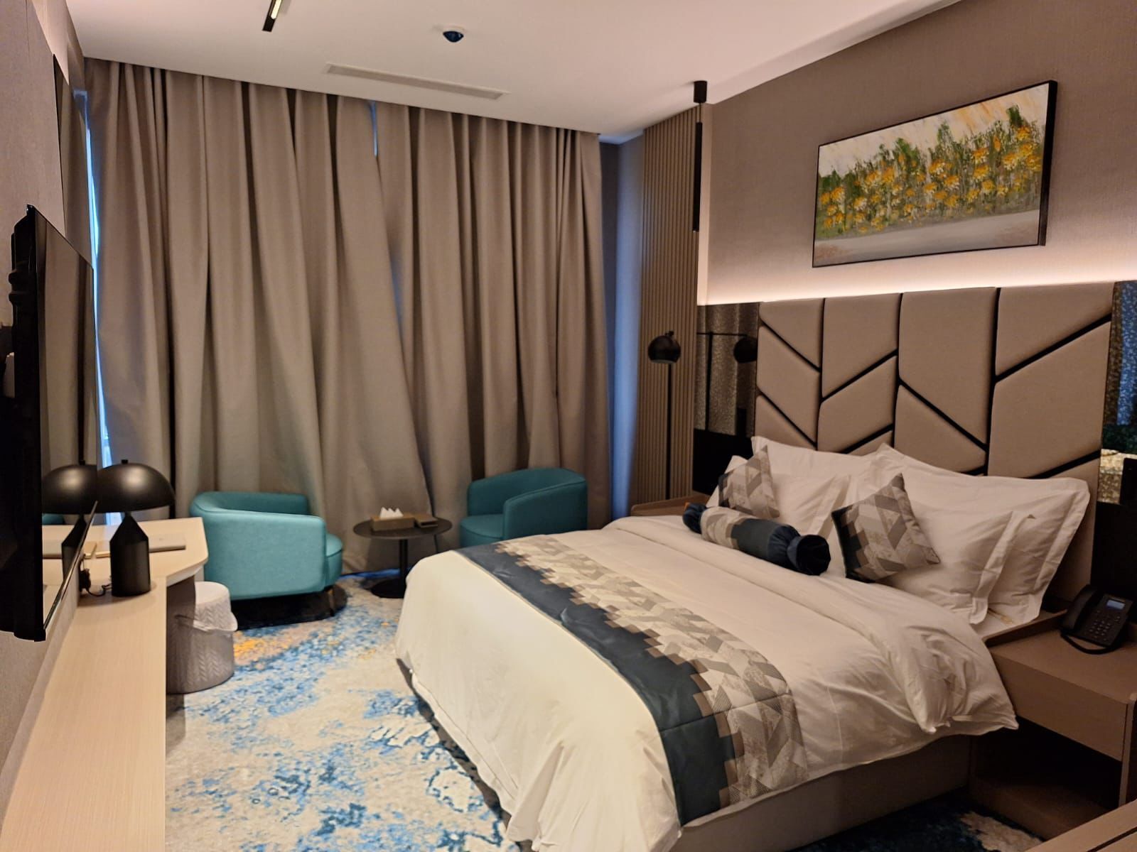 A modern hotel room with a large bed, two armchairs, a desk with a lamp, and a painting on the wall.