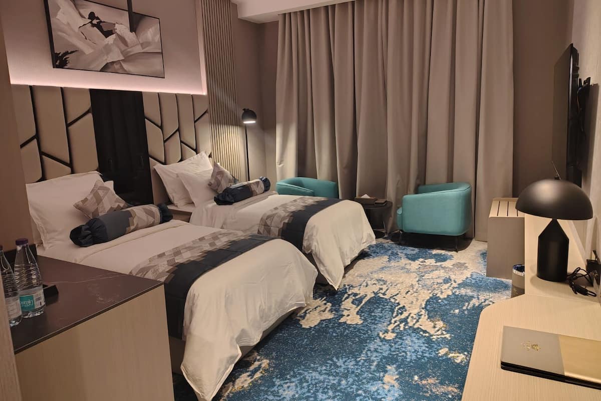 A hotel room with two beds, a desk, a chair, a TV, and a large window with curtains.