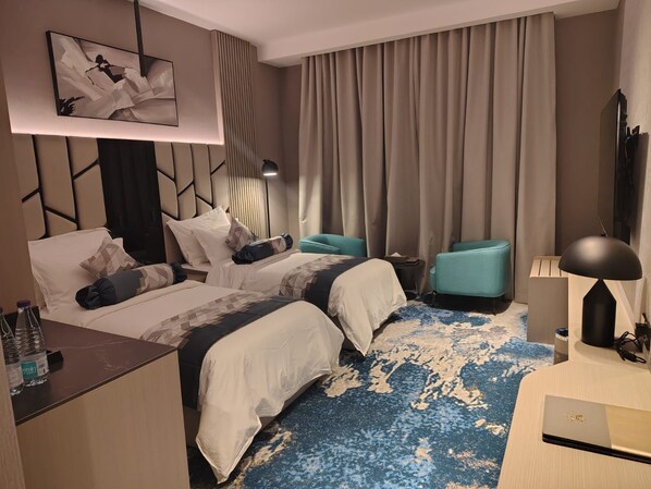 A hotel room with two beds, a desk, a chair, a TV, and a large window with curtains. - Taj Lives Hotel (Umluj)