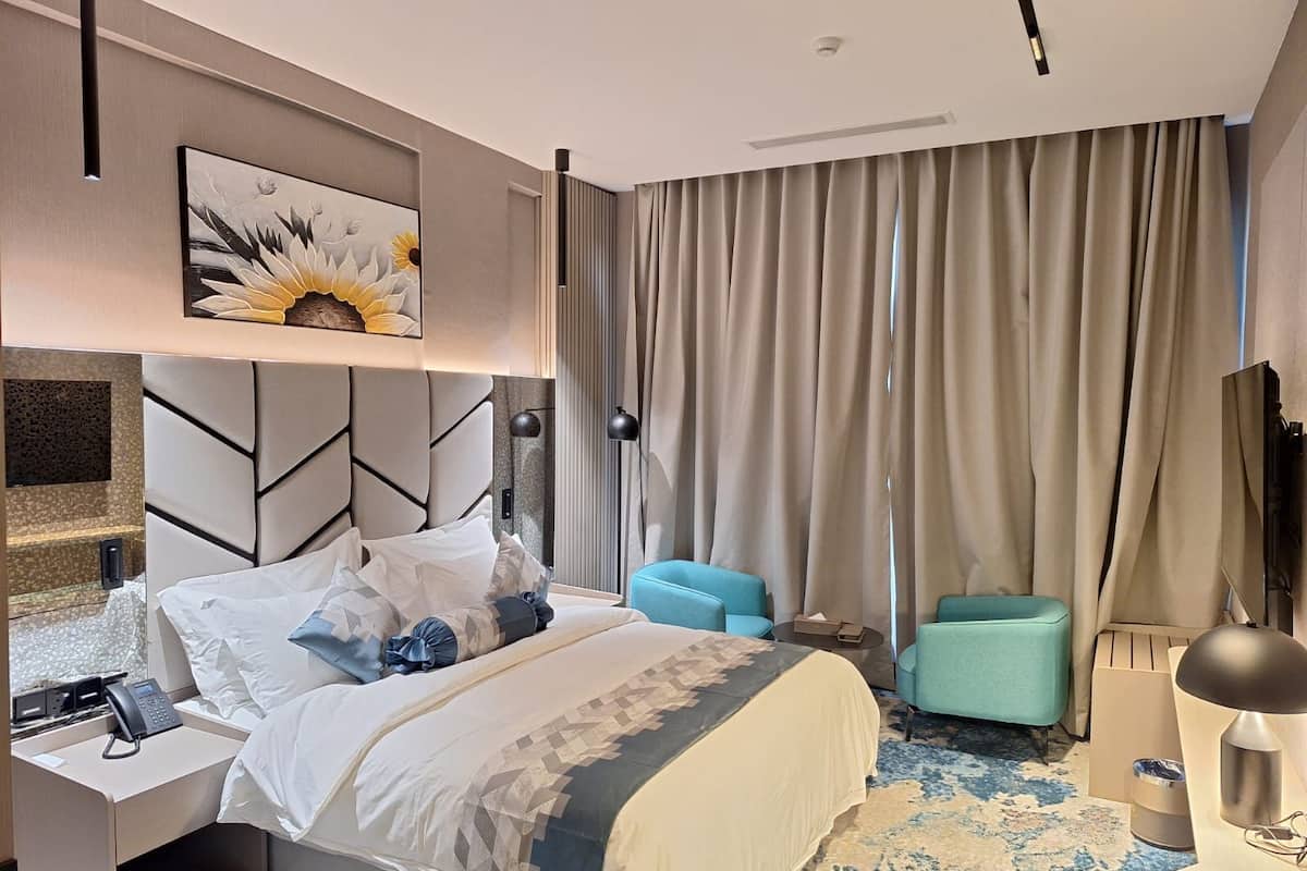 A modern hotel room with a large bed, a headboard with geometric patterns, a painting of sunflowers, a desk with a phone, a blue armchair, and a large window with curtains.