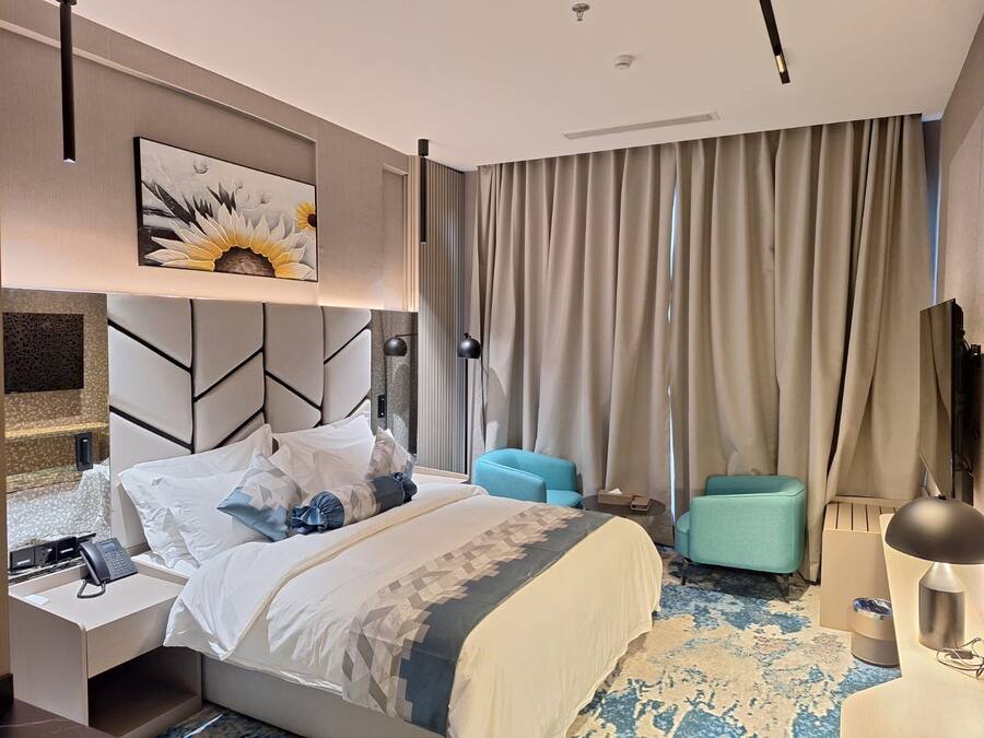 A modern hotel room with a large bed, a headboard with geometric patterns, a painting of sunflowers, a desk with a phone, a blue armchair, and a large window with curtains.