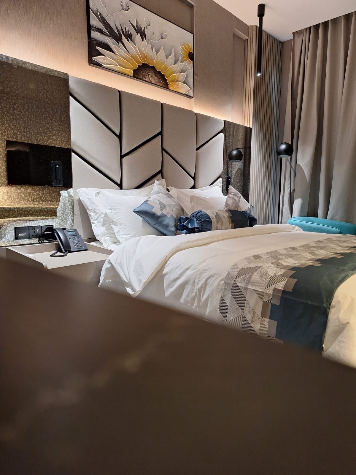 A modern hotel room with a large bed, a flat-screen TV mounted on the wall, a bedside table with a phone, and a decorative wall piece above the bed.
