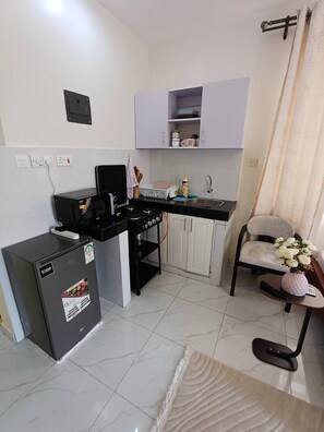 A compact kitchen with a refrigerator, stove, and sink. There are cabinets above the counter and a chair with a vase of flowers. - Vee’s Studio Airbnb in mtwapa (Mtwapa)