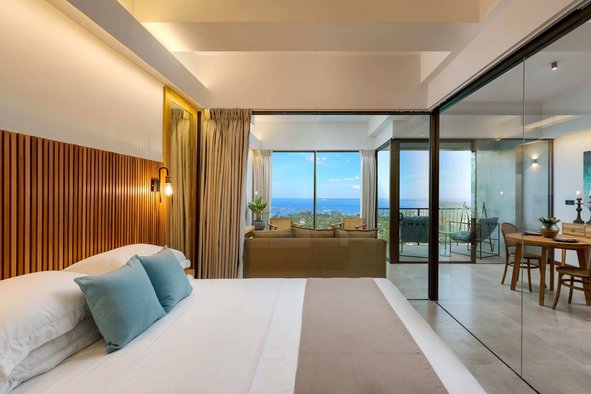 A modern hotel room with a large bed, a sofa, a dining area, and a balcony with a view.