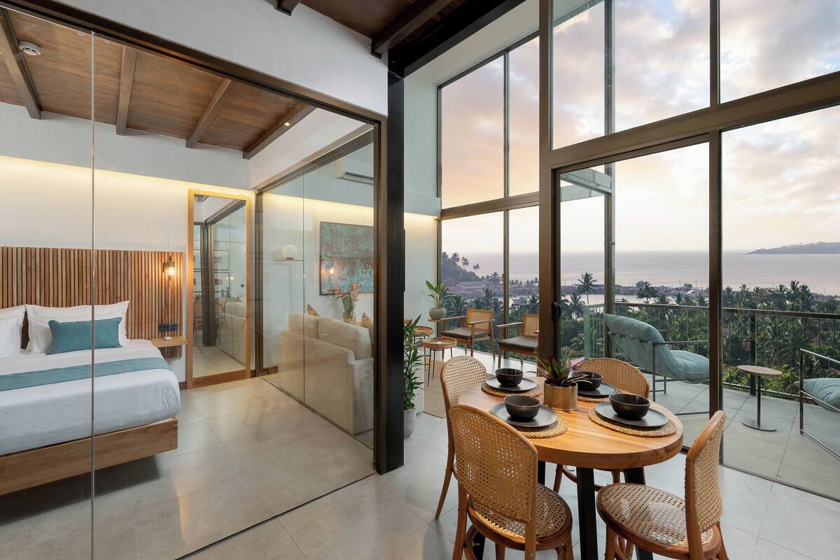 A modern room with a bed, a dining area with chairs and a table, and large windows offering a view of the ocean.