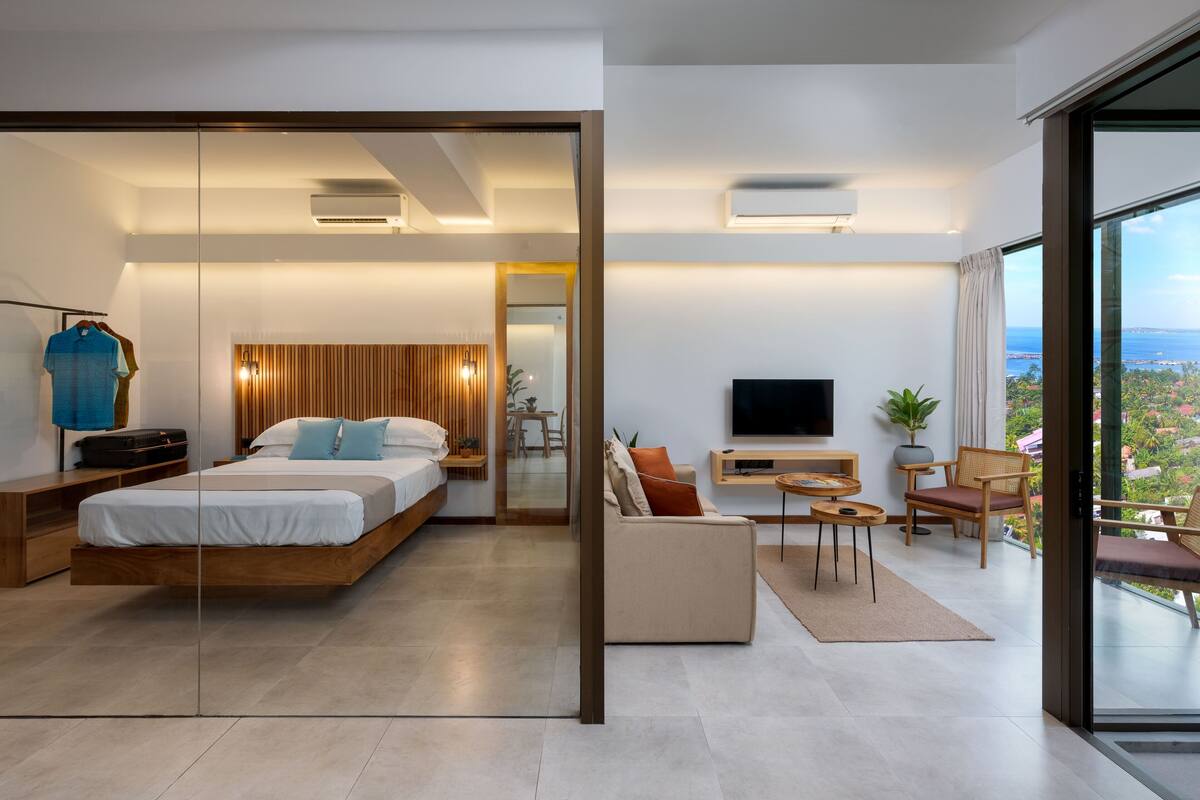 A modern hotel room with a bed, sofa, TV, and a balcony with a view.