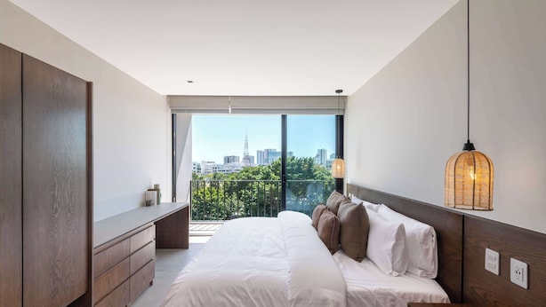 A modern bedroom with a large bed, a wooden dresser, and a city view through large windows. - Mojarra 27: Apt. with Balcony & King Bed (Cancún)
