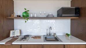 Eine moderne Küche mit weißer Arbeitsplatte, Spüle aus Edelstahl und einer Mikrowelle. Darüber befindet sich ein Holzregal mit einer grünen Vase, weißen Tassen und einem Stapel Teller.
