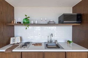 A modern kitchen with a white countertop, stainless steel sink, and a microwave. There is a wooden shelf above the counter holding a green vase, white mugs, and a stack of plates. - Mojarra 27: Cozy Apartment with Balcony (Cancún)