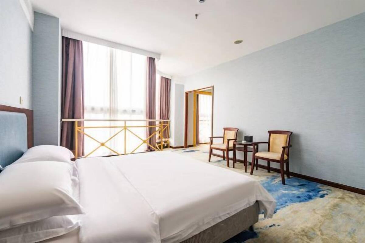 A hotel room with a bed, two chairs, a small table, and a large window with curtains.