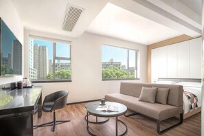 Ein modernes Hotelzimmer mit einer Couch, einem Couchtisch und einem Bett mit Kopfteil. Zwei Fenster bieten Blick auf die Stadt.