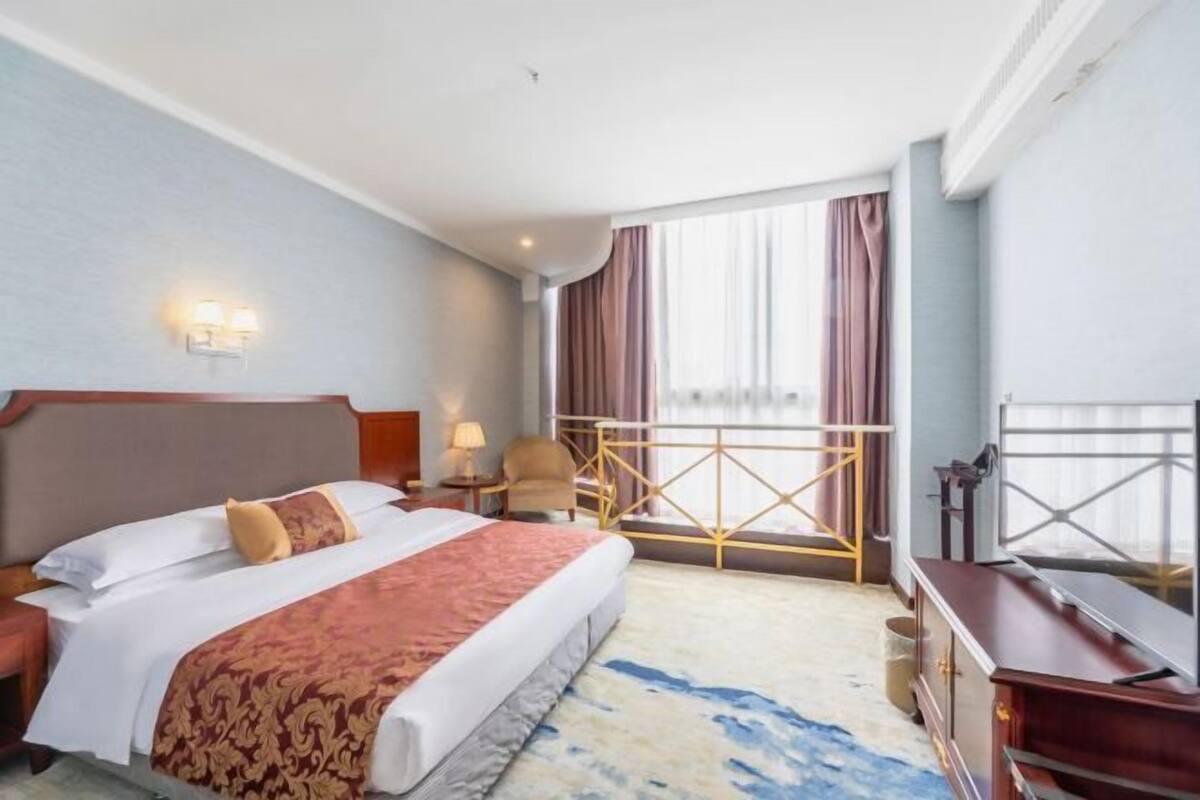 A hotel room with a large bed, a desk, a chair, a TV, and a balcony with curtains.