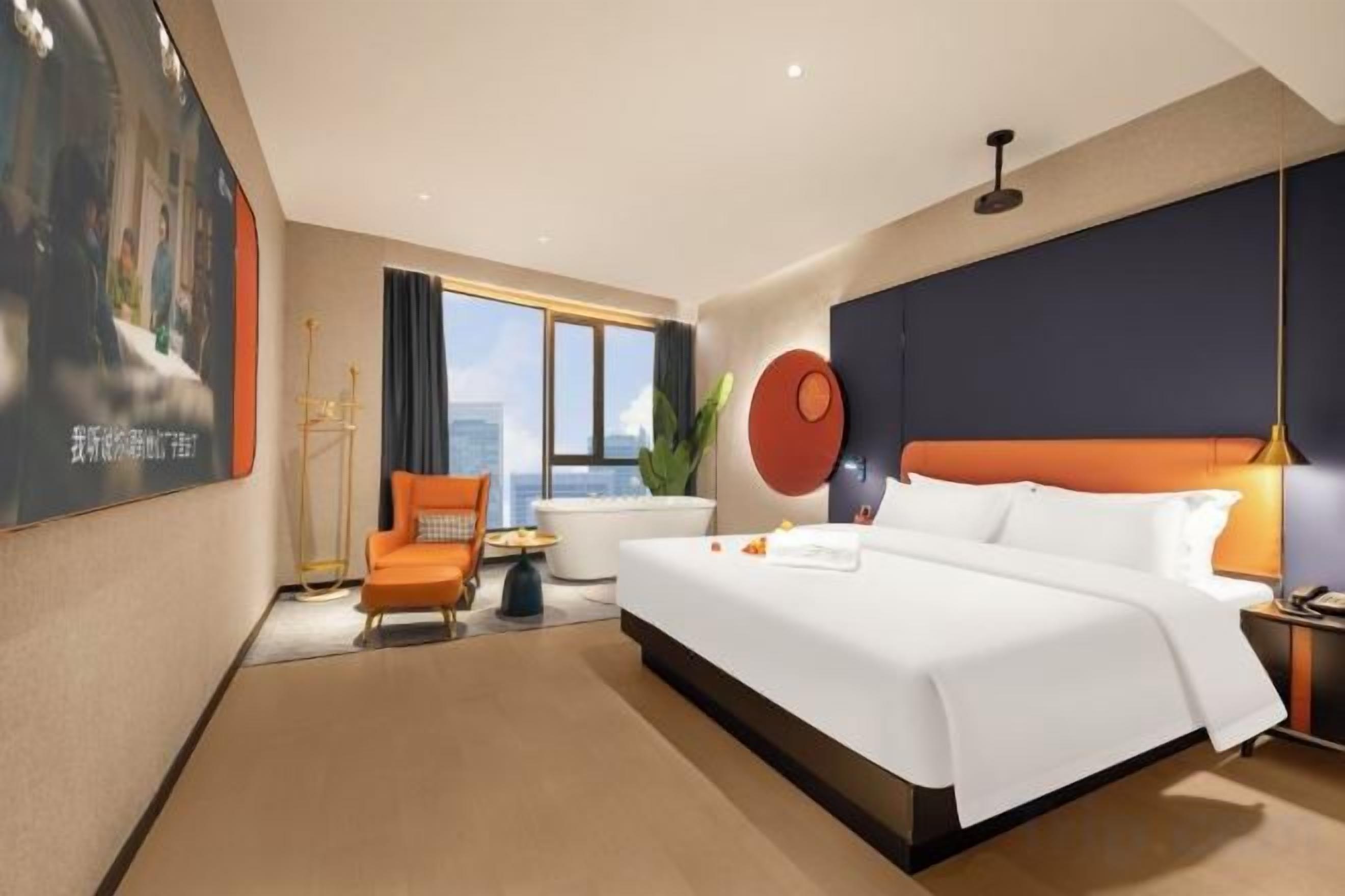 A hotel room with a large bed, a seating area with a chair, a window with curtains, and a wall-mounted artwork.