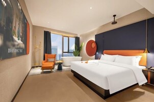 Habitación de hotel con una cama grande, zona de estar con una silla, ventana con cortinas y una obra de arte en la pared.
