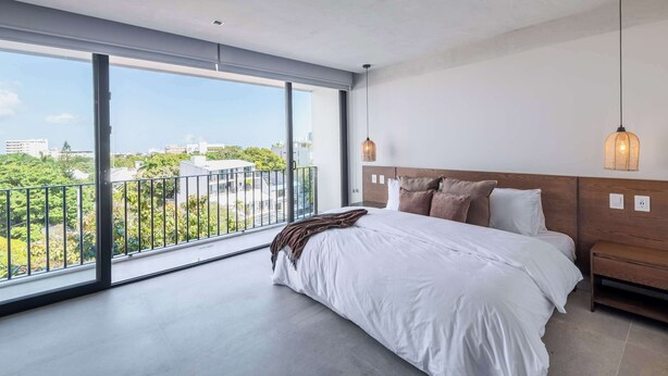 A modern bedroom with a large window, a bed with white bedding, a wooden headboard, and a hanging light fixture. - Mojarra 27: New Apt w/Balcony - Heart of Cancun (Cancún)