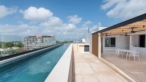 A rooftop pool with a clear view of the city skyline, a modern building with a glass facade, and a patio area with outdoor seating. - Mojarra 27: New Apt w/Balcony - Heart of Cancun (Cancún)