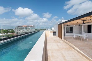 A rooftop pool with a clear view of the city skyline, a modern building with a glass facade, and a patio area with outdoor seating. - Mojarra 27: New Apt w/Balcony - Heart of Cancun (Cancún)