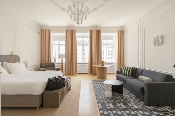 A hotel room with a bed, a sofa, a small table, and a view of buildings through large windows. - Nyhavn63 Premium Apartment 1 Daniel&Jacob's (København)