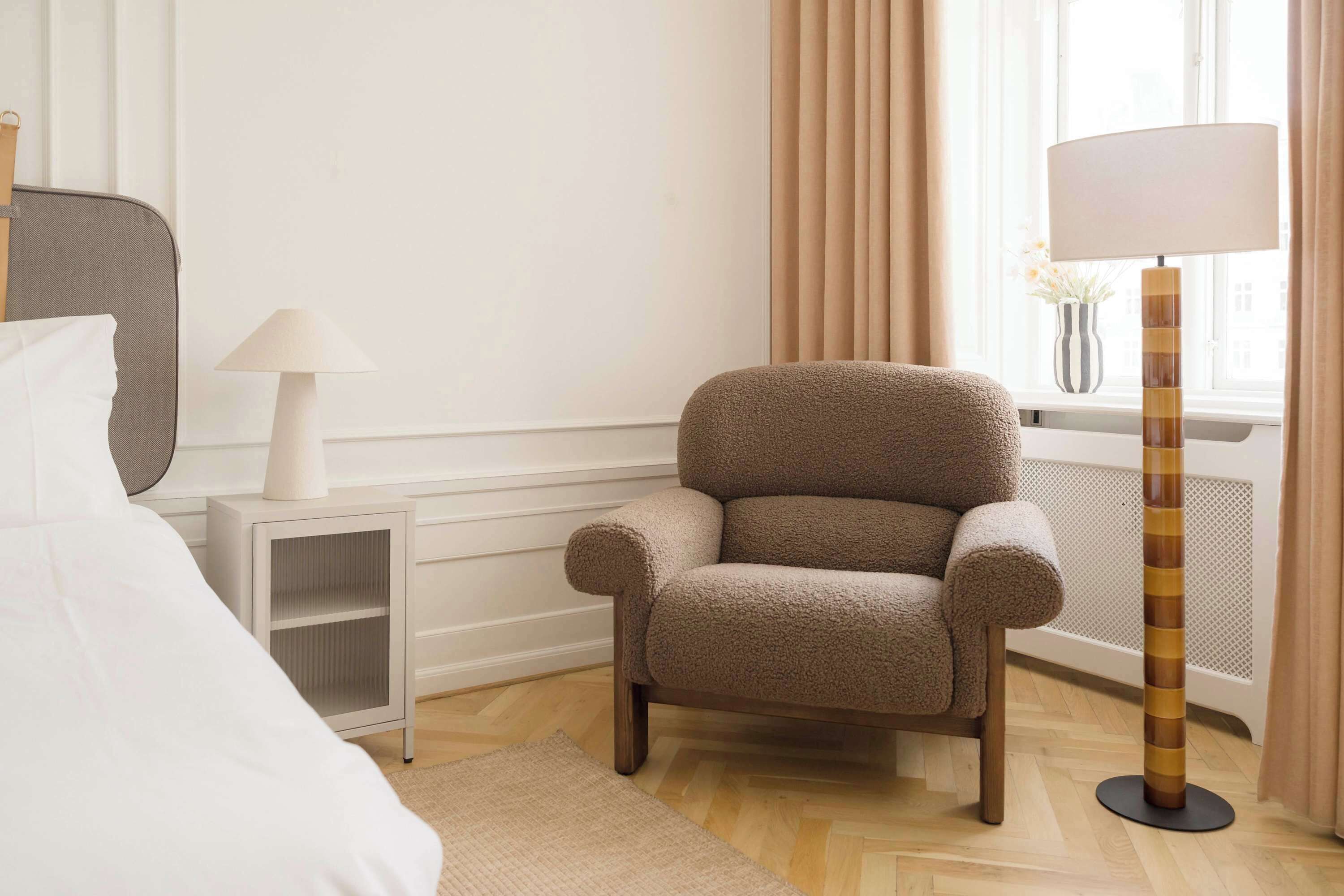 A hotel room with a bed, a chair, a lamp, a floor lamp, and a window with curtains.