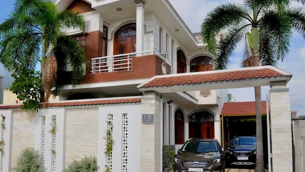 Una casa a due piani con un balcone, un garage con due automobili e palme.