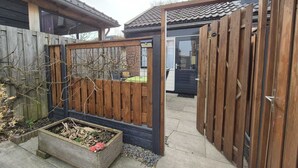 Una cerca de madera con un portón, una jardinera de madera con plantas, y un tejado de baldosas visible al fondo.