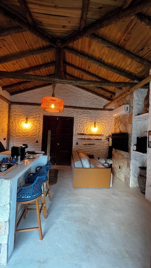 A rustic interior with a stone wall, a wooden ceiling, a sofa, a bar area with stools, and a television mounted on the wall.