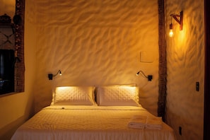 A neatly made bed with a textured wall behind it, two wall-mounted light fixtures, and a small wall switch.