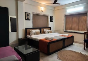 A bedroom with a bed, bedside tables, a glass table, a ceiling fan, and a window with blinds. - The Kolam (Bolpur)