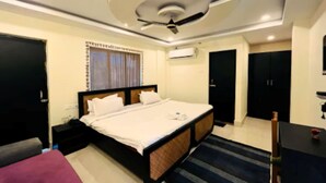 A hotel room with a bed, a desk, a chair, a ceiling fan, and a window with curtains. - The Kolam (Bolpur)